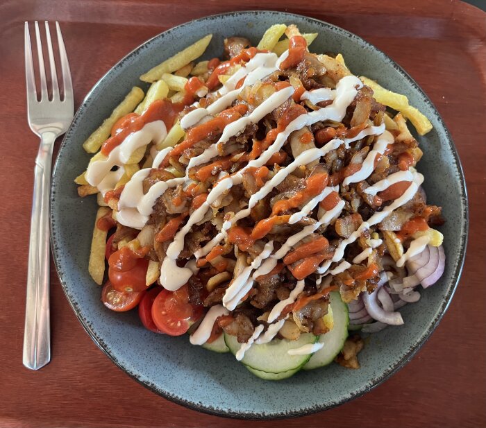 Pommes frites med kebabkött, såser, grönsaker på en grå tallrik och gaffel på bordet.