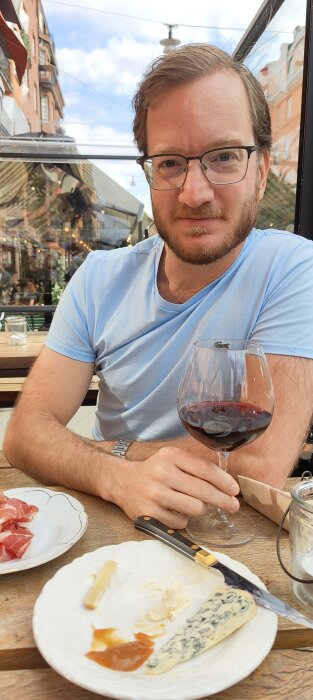 Man sitter utomhus, dricker vin, äter ostar och charkuterier; stadsbakgrund, avslappnad atmosfär.