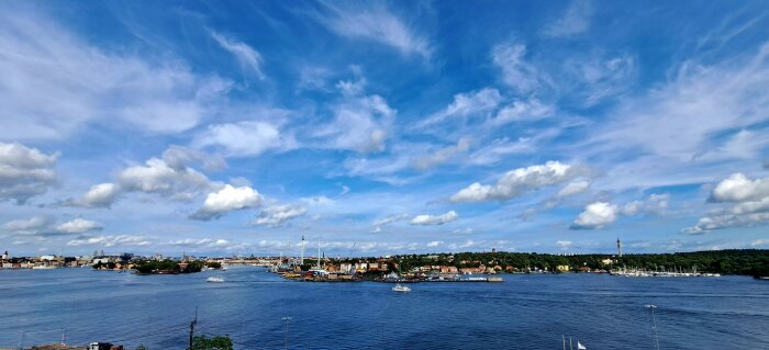 Stadspanorama, vatten, båtar, moln, klar himmel, gröna träd, byggnader, kulturlandskap.