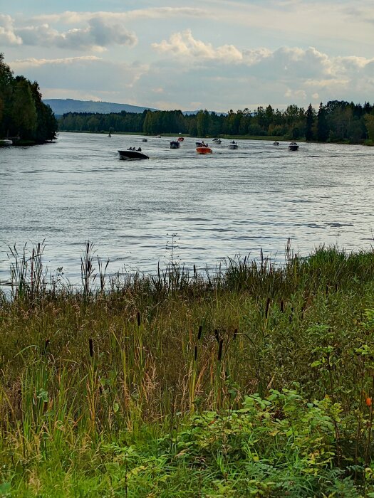 Båtar på en flod, grönska, molnig himmel, natur, rekreation, vegetation i förgrunden, kuperad terräng i bakgrunden.