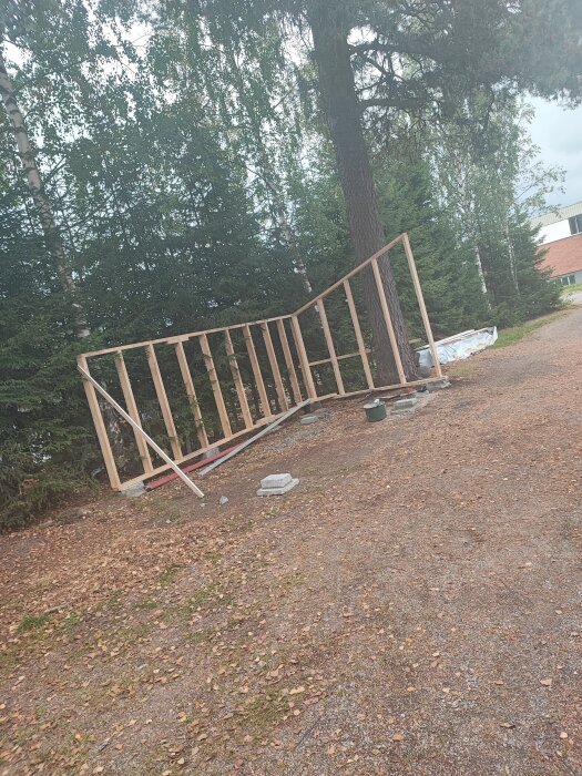 Stort trästaket ligger på marken vid en grusväg omgiven av träd, himmel synlig, konstruktionsmaterial runtomkring.