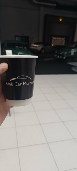 Hand håller pappersmugg med "Saab Car Museum"-tryck, bilar i bakgrunden i ett museum.