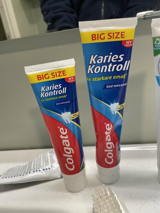Två stora tandkrämstubar och en tandborste på badrumsbänk. "Karies Kontroll", Colgate, mintsmak.