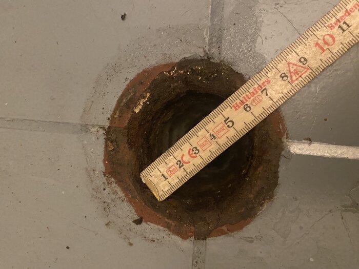 Ett måttband som sträcker sig över ett cirkulärt hål i betong, synlig jord, konstruktion eller reparationsarbete pågår.