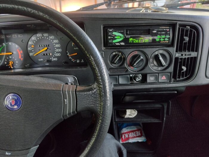 Bild på en Saab-bils interiör med ratt, instrumentpanel, stereo och öppnat handskfack med saker i.