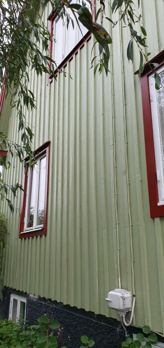 Grönmålad plåtfasad, fönster, röda fönsterkarmar, växtlighet, elutrustning, del av ett hus.