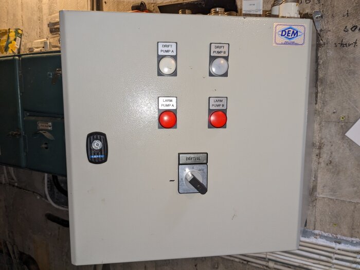 Elektrisk kontrollpanel med knappar och indikatorlampor för pumpar, larm och temostat, placerad i ett industriellt utrymme.