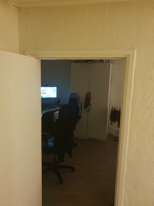Enkelt arbetsrum med dator, skrivbordsstol, garderob och kläder hängande på en stol.