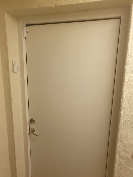 Vit dörr med dörrhandtag, lås och intercom panel i en vägg med grov struktur.