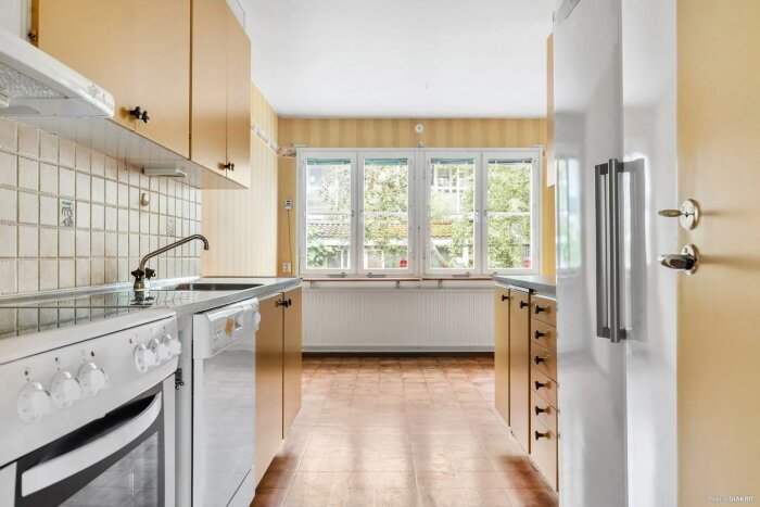Ett ljust kök med trämöbler, kakelvägg, spis, kylskåp och fönster med utsikt mot grönska.