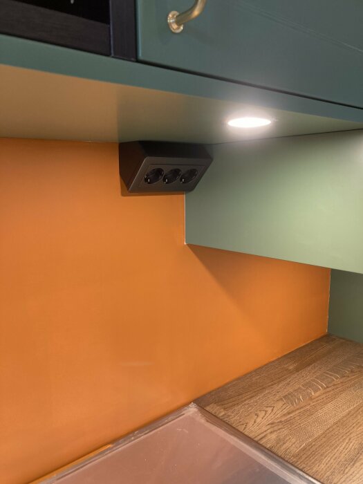Inbyggd spotlight och eluttag under hylla, modern köksdesign, gröna och orangea toner, trägolv.