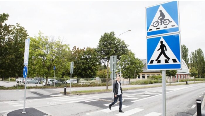 Man korsar gata vid övergångsställe, trafikskyltar för gående och cyklister, träd och parkerade bilar i bakgrunden.