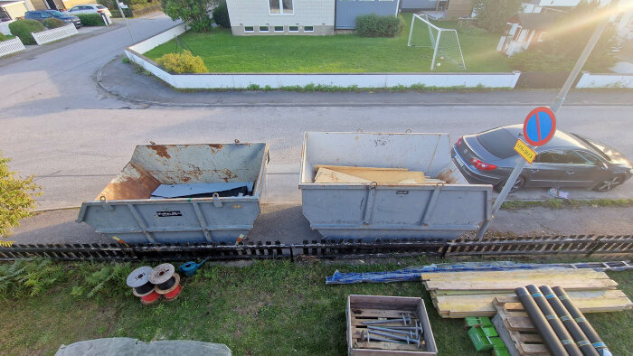 Två stora container, byggmaterial, parkerad bil, bostadshus, grön gräsmatta, fotbollsmål, vägskylt, tyst villakvarter.