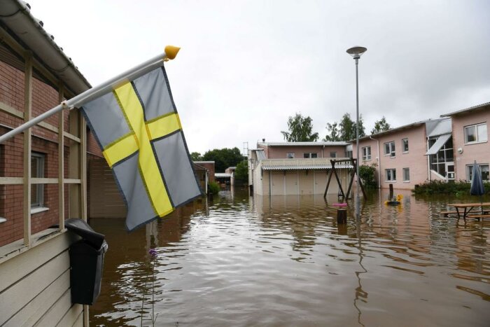 Svensk flagga, översvämmad bostadsområde, vatten upp till husens första våning, molnig himmel, ingen synlig person.