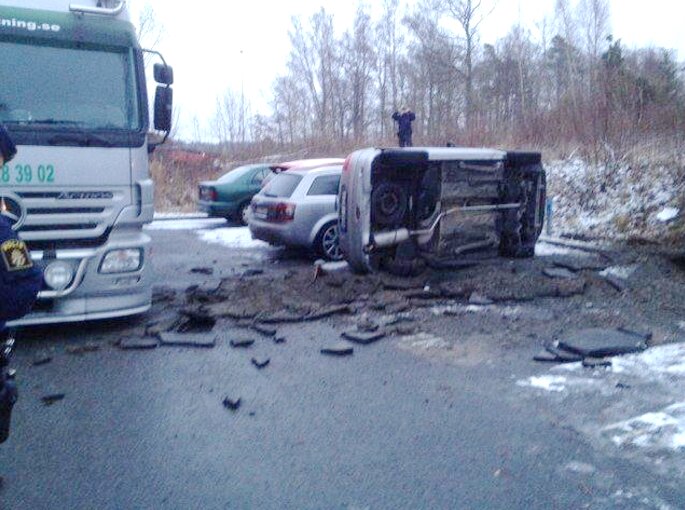 Trafikolycka med en personbil som ligger på sidan på en väg, omgiven av spillror och en lastbil intill.