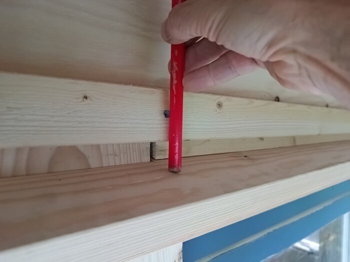 Hand håller röd penna mot träbjälkar för mätning eller markering vid byggprojekt.