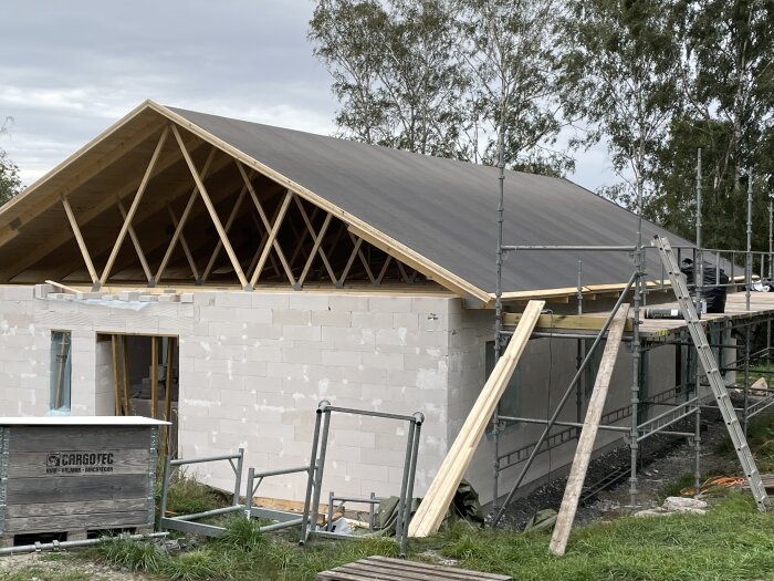 Byggarbetsplats med halvfärdigt hus, synliga takstolar, byggställningar och byggmaterial på marken.