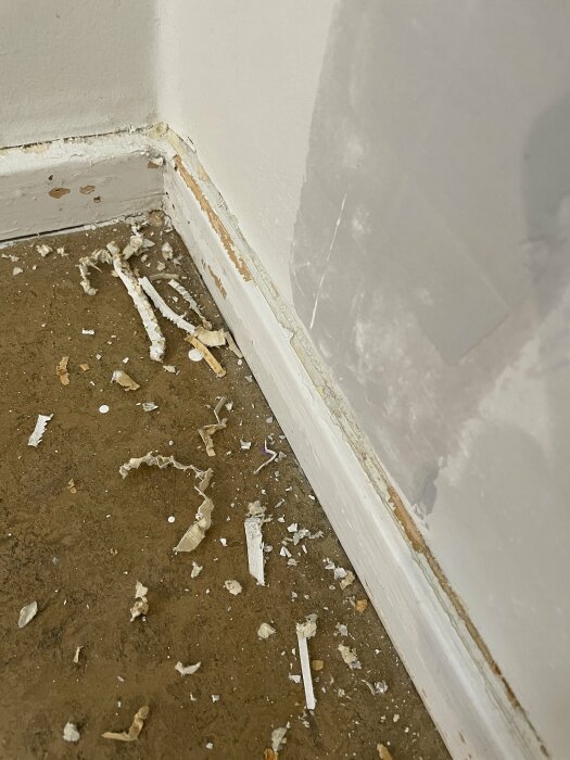 Hörn av ett rum med skadade lister, trasigt material på smutsigt golvet. Renovering eller skada synlig.