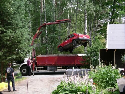 Röd bil lyfts av kran till lastbilsflak i skogsområde medan personer observerar.