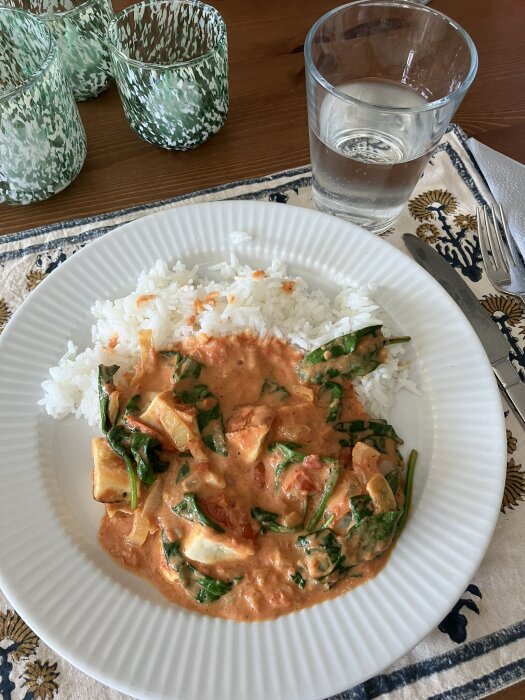 Tallrik med ris, tomat- och paneer gryta, spenat, vattenglas, bestick, mönstrad duk.