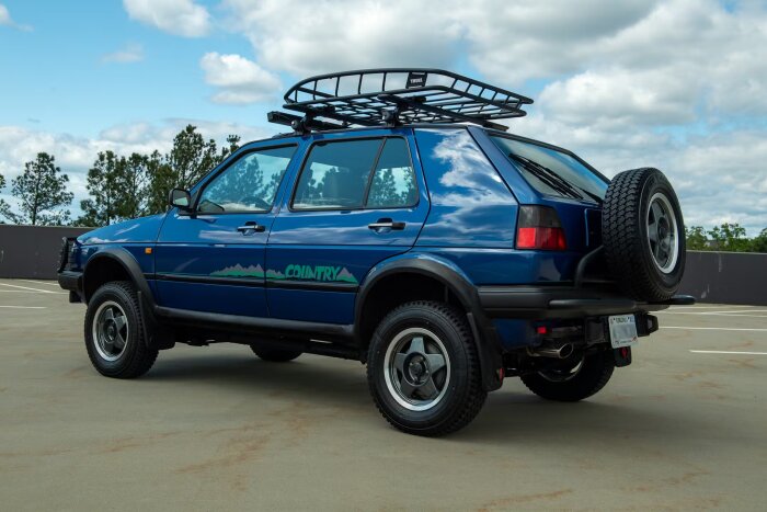Blå SUV med takräcke och reservhjul bak, parkerad på en asfalterad yta.