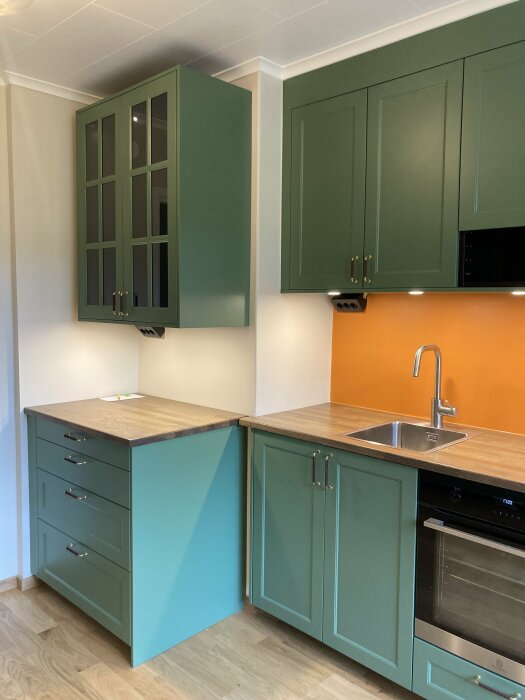 Ett modernt kök med gröna skåp, träbänkskiva, inbyggd ugn och orange stänkskydd.