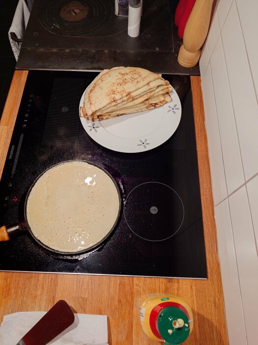 Tillagning av pannkakor på spishäll, stapel på tallrik, syltburk och redskap närvarande. Hygienpapper vid sidan.