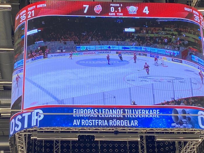 Ishockeymatch visas på stor skärm, period 3, hemmalag leder med 7-4, publiken synlig.