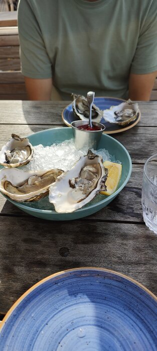 Ostron på is med citron och sås, person i grönt, uteservering, blå tallrik, träbord, vattenglas.