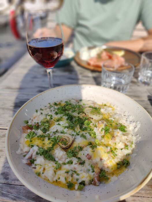 Risrätt med persilja, citronskiva, rostad lök; rödvin, andras maträtter i bakgrunden; utomhusmiddag.