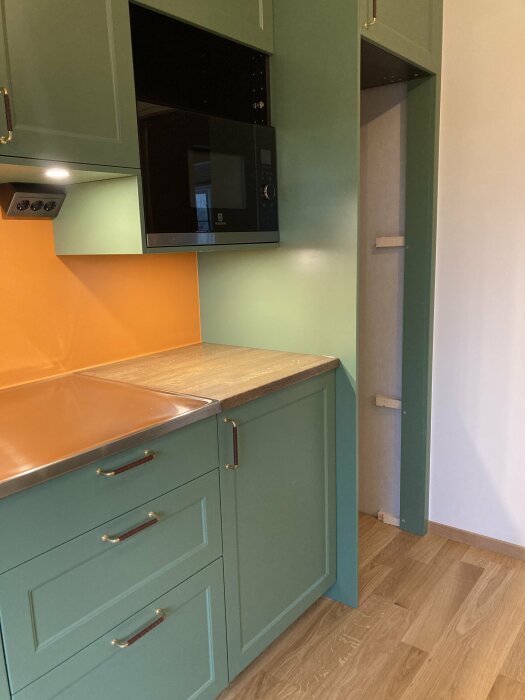 Modernt kök, gröna skåp, inbyggd mikrovågsugn, trägolv, orange bänkskiva, undanskymd kylskåpsdörr.