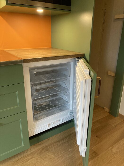 Kök med öppen, tom frys, gröna skåp, träbänkskiva och orange vägg.