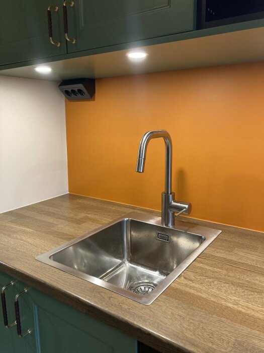 Modernt kök, rostfri diskho, orange vägg, gröna skåp, belysning under överskåp, träarbetsbänk.