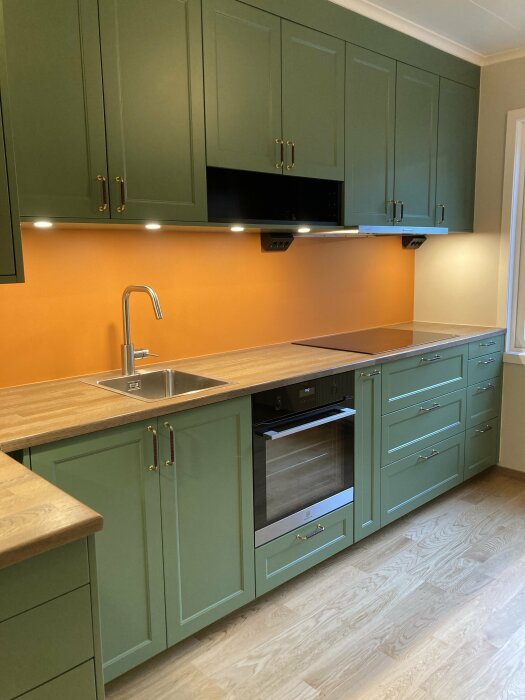 Modernt kök, gröna skåp, träbänkskiva, inbyggd ugn, LED-belysning under skåp, rostfri vask och vitt golv.