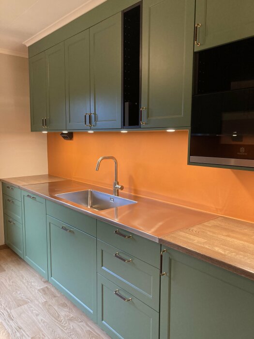 Modernt kök med gröna skåp, rostfri diskho, mikrovågsugn, träbänkskiva och orange stänkskydd.