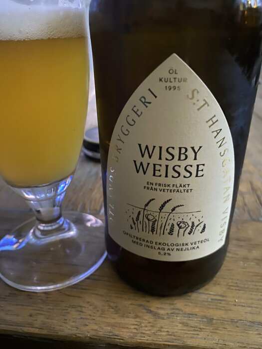 Ölflaska med etikett "Wisby Weisse" bredvid ett glas med öl, på ett träbord.