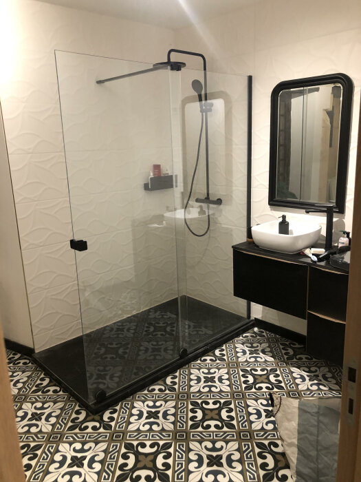 Modernt badrum, duschhörna med glasvägg, mönstrat golv, handfat, spegel, svarta och vita detaljer.