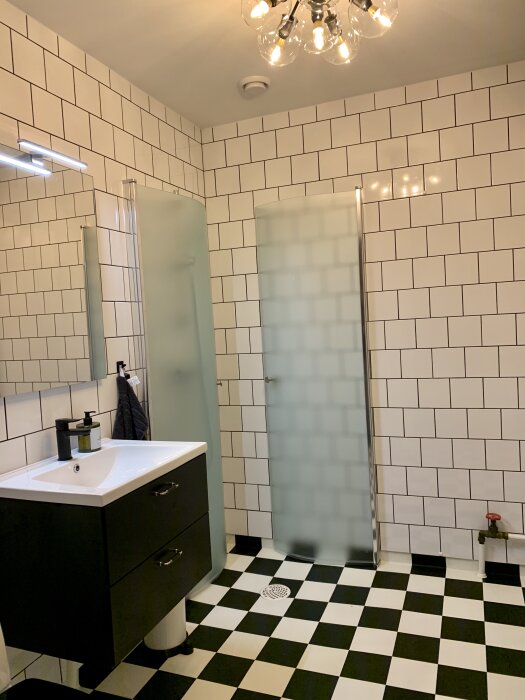 Badrum med svartvit rutigt golv, vit kakelvägg, duschhörna, handfat och spegel.
