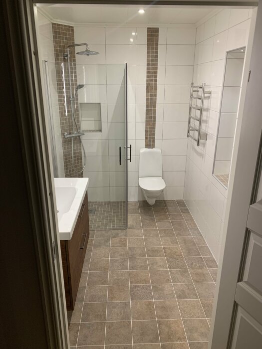 Modernt badrum med dusch, toalett, handfat, kakel, handdukstork och brunt golv.