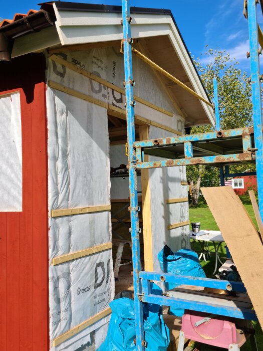 Byggnadsställning vid hus under renovering, isoleringsmaterial synligt, solljus, trädgård i bakgrunden.