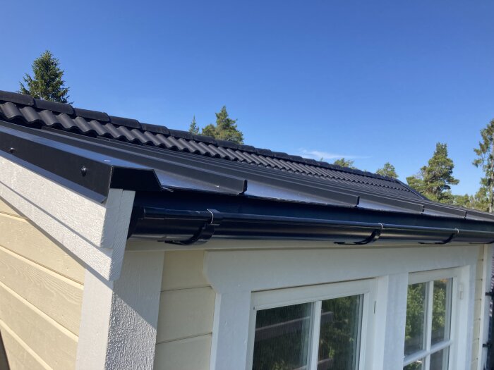 House corner, black tiled roof, gutter, sunny day, trees in background, clear blue sky.