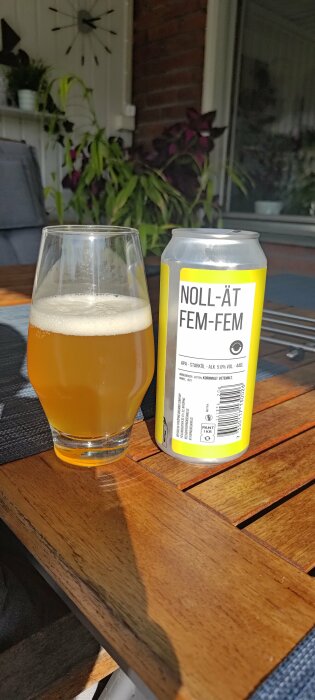Ett glas öl och en burk med texten "NOLL-ÄT FEM-FEM" på ett träbord utomhus.