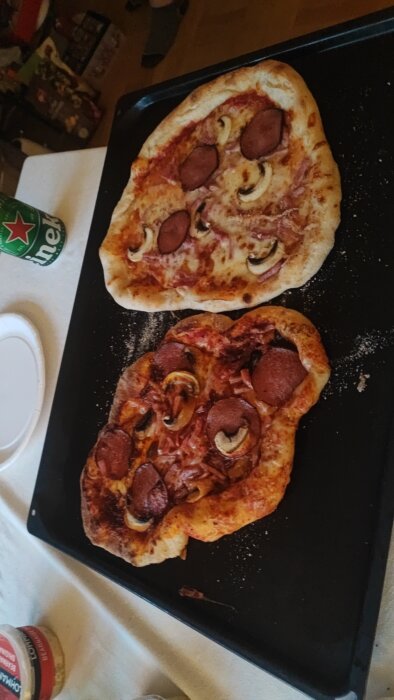 Två hembakade pizzor med pepperoni och svamp på bakplåt, vardagsröra i bakgrunden.