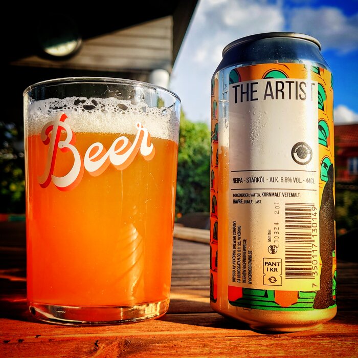 Ölglas märkt "Beer" fyllt med öl, bredvid burk märkt "THE ARTIST", solljus, träyta.