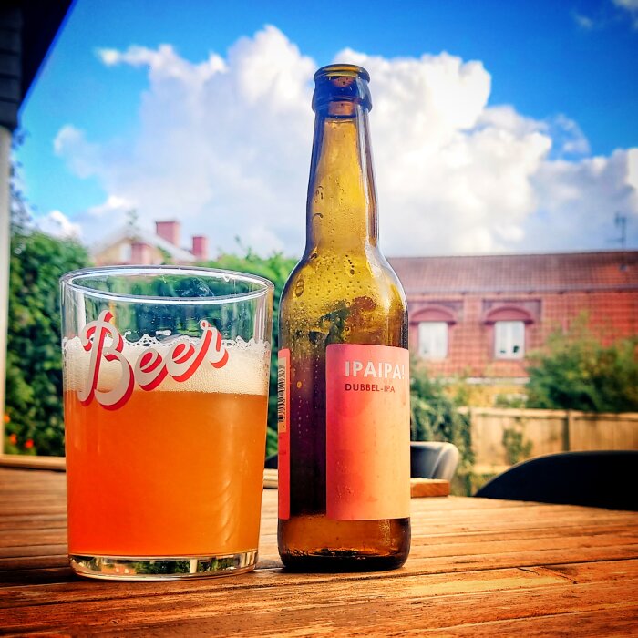 Ölglas och flaska på träbord, utomhus, blå himmel, vita moln, byggnader i bakgrunden.