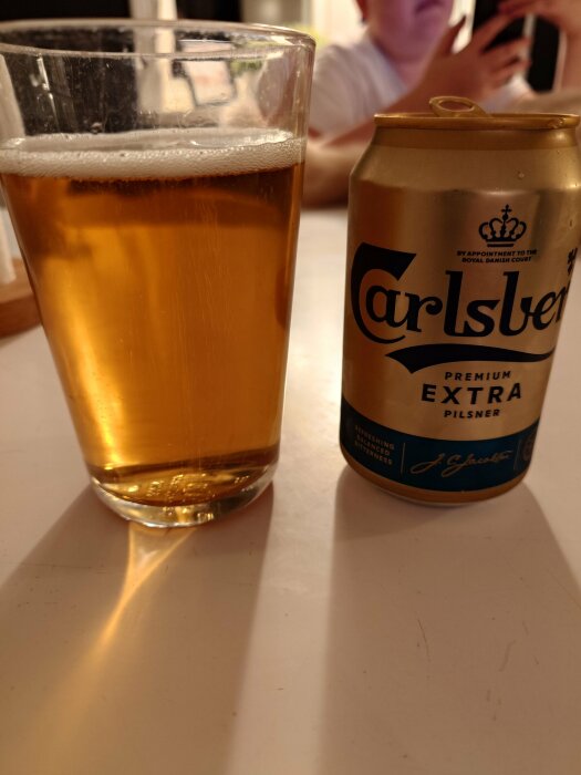 Ett glas öl bredvid en öppnad Carlsberg burk, inomhus, möjlig social samvaro.