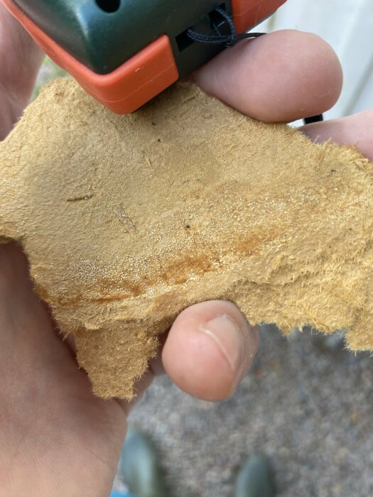 Hand håller ett stycke grovt tyg eller filt med grön och orange plastdel ovanför.