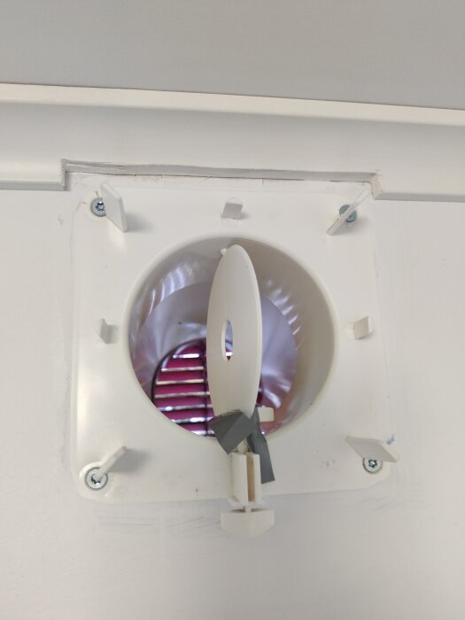 Ventilationsöppning med vit ram och delvis öppen spjäll, fastsatt i ett tak, tejpbitar synliga.