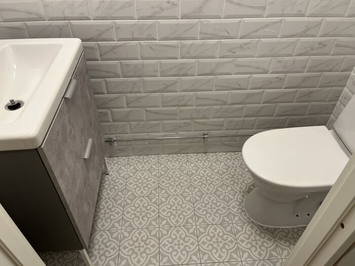 Ett badrum med handfat, toalett, kakelväggar och mönstrat golv. Modernt och rent intryck.