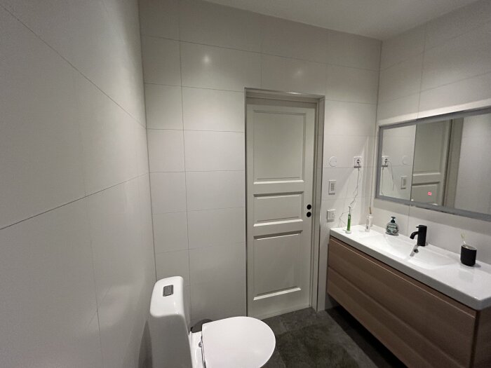 Modernt badrum med vit toalett, handfat, spegel, gråa kakelväggar och brun skåpinredning.